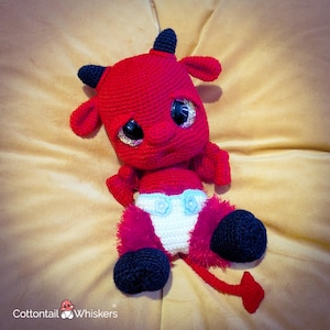 Pode incluir: Boneca de diabo vermelha de crochê feita à mão com chifres, orelhas e pés pretos. A boneca tem olhos grandes e brilhantes e uma fralda branca com detalhes de flores azuis. Uma cauda vermelha se estende da parte de trás da boneca.