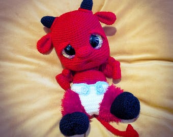 May include: A handmade red crochet devil doll with black horns, ears, and feet. The doll has large, sparkly eyes and a white diaper with blue flower accents. A red tail extends from the doll's back. The doll is sitting on a tan surface.