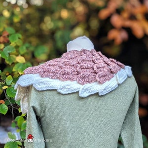 Pu&ograve; includere: Un collo in crochet rosa e bianco con un motivo a forma di squame. Il collo &egrave; indossato su un manichino con un maglione verde.