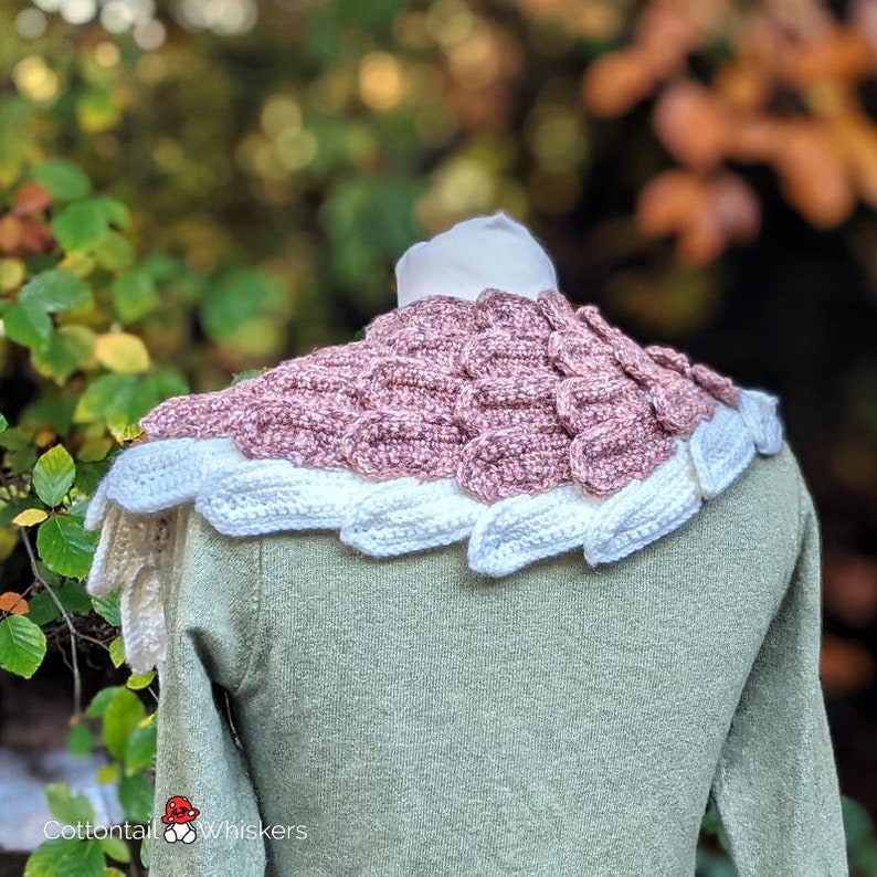 Pu&ograve; includere: Un collo in crochet rosa e bianco con un motivo a forma di squame. Il collo &egrave; indossato su un manichino con un maglione verde.