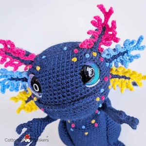 May include: A handmade, crocheted axolotl plushie in shades of navy blue, with colorful accents. The plushie features bright pink, blue, and yellow gills, and small, colorful dots. The eyes are large and glittery, with a black pupil and a blue iris.
