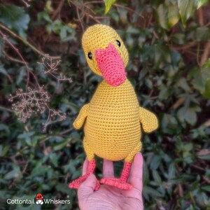 May include: A handmade yellow crochet duck with a bright pink beak and feet. The duck has small black eyes and is held in a person's hand. The background features green foliage and the logo "Cottontail Whiskers".