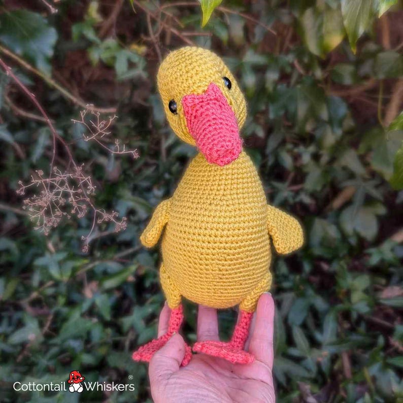 May include: A handmade yellow crochet duck with a bright pink beak and feet. The duck has small black eyes and is held in a person's hand. The background features green foliage and the logo "Cottontail Whiskers".