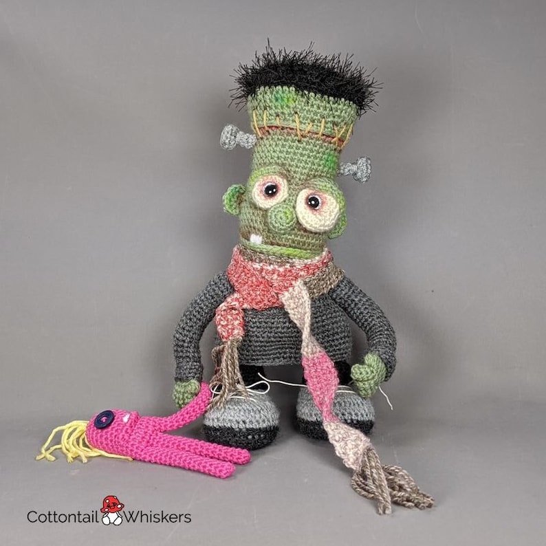 Pode incluir: Um boneco de Frankenstein de croch&ecirc; verde vestindo um su&eacute;ter cinza e uma echarpe rosa e cinza. O boneco est&aacute; de p&eacute; com os bra&ccedil;os estendidos e tem um sorriso costurado. Um boneco de croch&ecirc; rosa com cabelo amarelo est&aacute; deitado no ch&atilde;o na frente do boneco monstro.