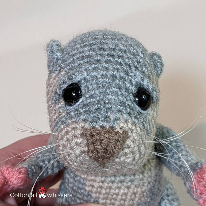 May include: A crocheted gray and brown otter with black eyes and a pink nose. The otter has long white whiskers.