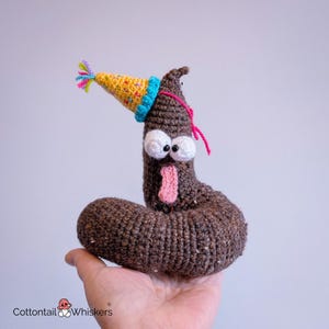 May include: A brown crocheted plush toy shaped like a coiled figure with a silly expression, featuring large white eyes, a pink tongue, and a yellow party hat with colorful speckles and a multi-colored tassel on top, held in a palm.