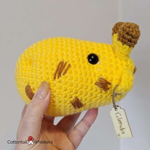 Puede incluir: Un animal de peluche de color amarillo tejido a crochet con detalles bordados marrones y un ojo negro. El animal de peluche tiene una peque&ntilde;a etiqueta de madera adjunta que dice "Glenda".