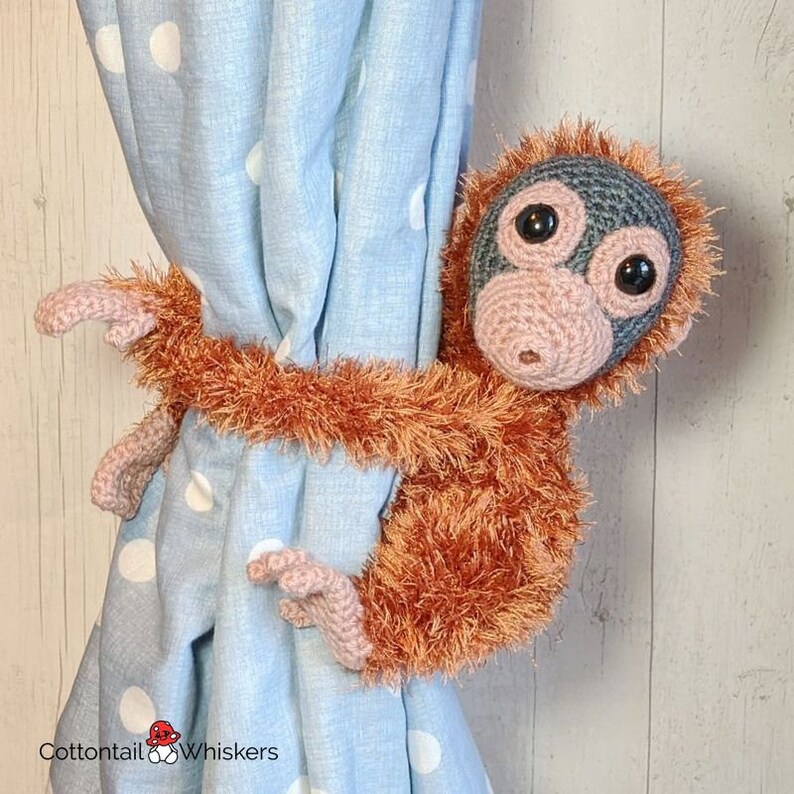 May include: A crocheted orangutan holding a blue and white polka dot curtain. The orangutan is brown with a gray face and large black eyes. The orangutan is holding the curtain with its arms and legs. The text "Cottontail Whiskers" is visible in the bottom left corner of the image.