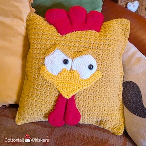 Peut inclure: Un coussin carré en crochet jaune moutarde représentant un visage de poulet. Le coussin présente une crête rouge en crochet sur le dessus, deux grands yeux blancs avec des pupilles noires, un bec jaune et deux barbillons rouges. Le texte Cottontail Whiskers est visible.