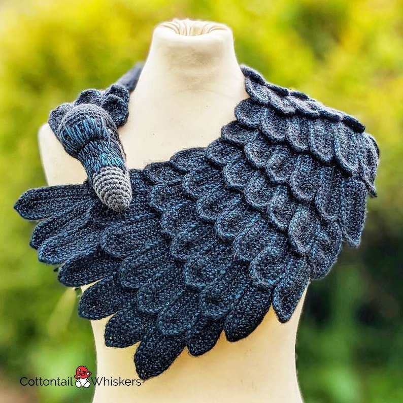 May include: A crocheted blue and black wing-shaped scarf with a small crocheted bird attached. The scarf is displayed on a white mannequin.