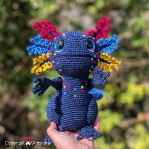 May include: A handmade, dark blue crochet axolotl plushie with red, yellow, and blue accents. The axolotl has large, black eyes and colorful gills. The plushie is held in a person's hand.