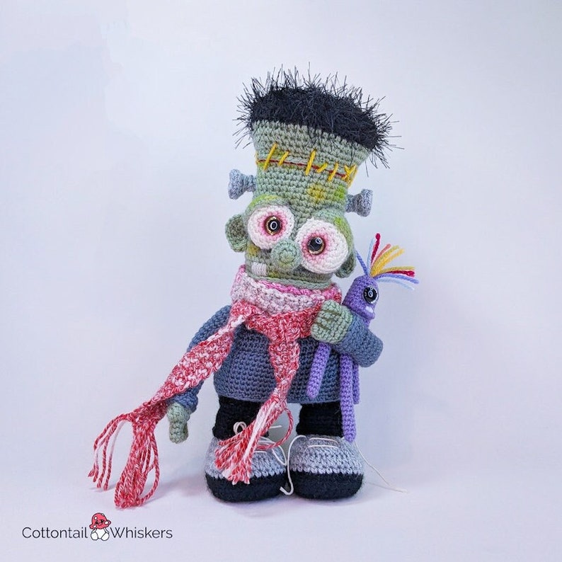 Pode incluir: Um monstro Frankenstein de croch&ecirc; verde com cabelo preto, usando uma echarpe vermelha e branca e um su&eacute;ter cinza, segurando um monstro de croch&ecirc; roxo com cabelo arco-&iacute;ris.