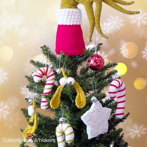 Könnte beinhalten: Ein Weihnachtsbaum, der mit handgefertigten Häkelornamenten geschmückt ist. Die Baumspitze ist ein grüner Grinch-Arm und ein rot-weißer Strumpf. Zu den Ornamenten gehören Zuckerstangen, ein Stern und Mistelzweige. Der Hintergrund ist goldfarben mit Schneeflocken.