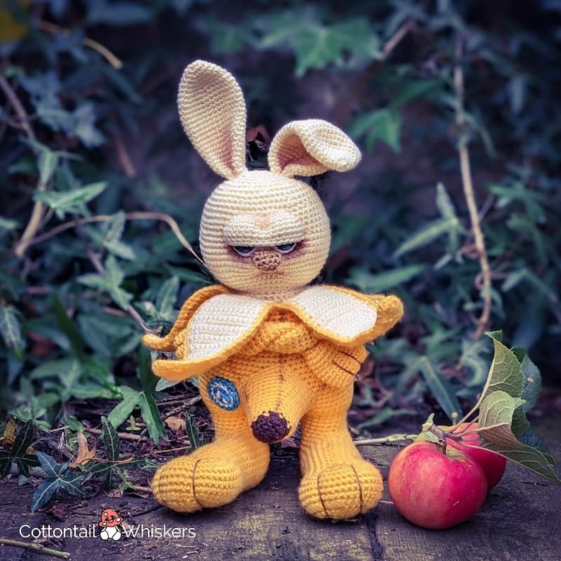 May include: A crocheted stuffed bunny wearing a banana costume. The bunny has a yellow body, white ears, and a brown nose. The banana costume is yellow and has a brown stem. The bunny is sitting on a wooden surface with a red apple in the background.