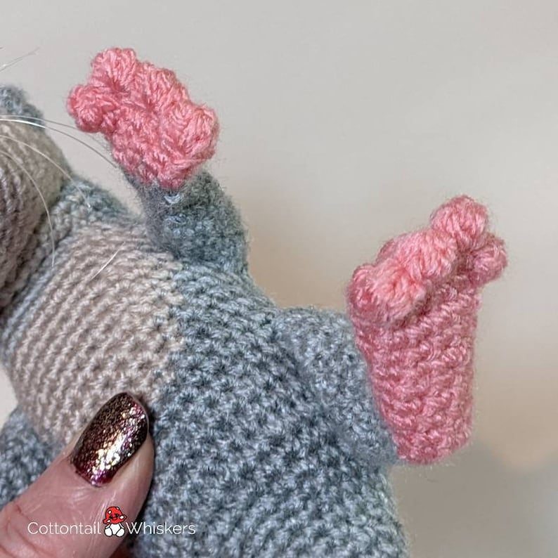 May include: Close-up of a crocheted gray and beige mouse with pink feet. The mouse is made with a textured stitch and has a soft, cuddly appearance.