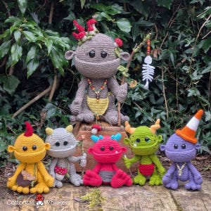 Könnte beinhalten: Eine Sammlung handgefertigter Häkelmonster-Puppen in verschiedenen Farben. Die Puppen haben einzigartige Merkmale, darunter Hörner, Hüte und Accessoires. Die Puppen werden vor einem grünen Hintergrund präsentiert. Die Puppen sind zwischen 15 und 30 cm groß.