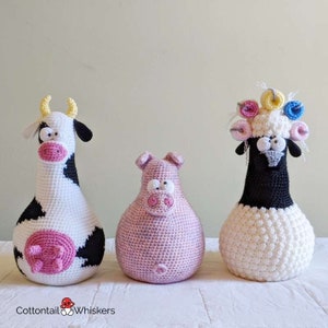 Op de afbeelding: Drie gehaakte knuffels: een koe met zwarte en witte vlekken, een roze varken en een wit schaap met een zwart gezicht en een kleurrijke bloemenkrans.