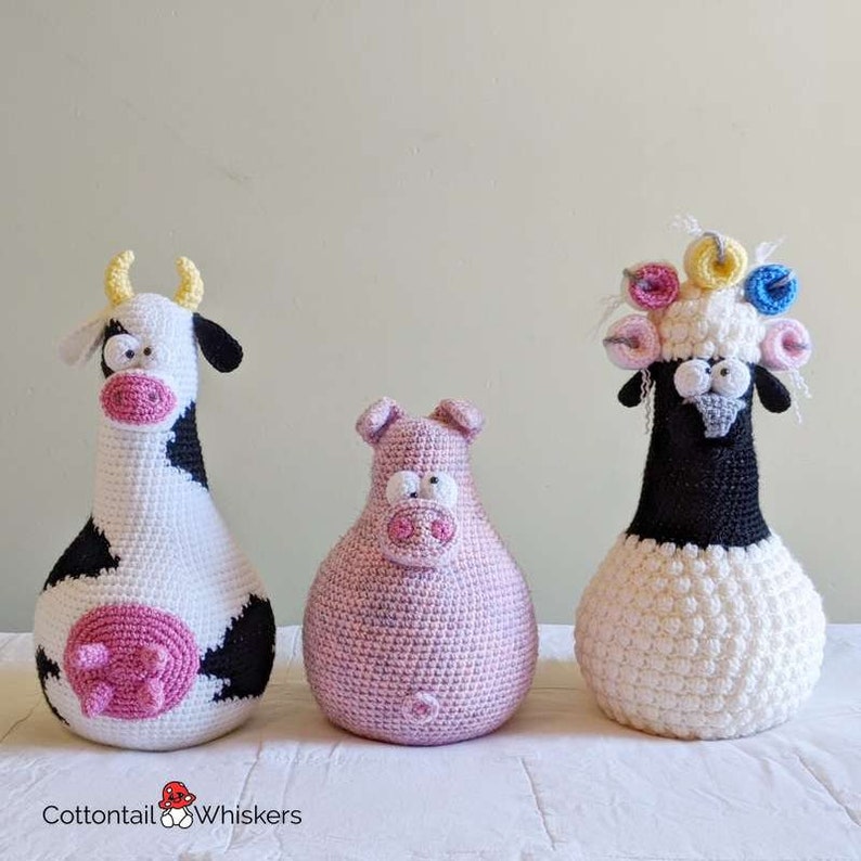 Op de afbeelding: Drie gehaakte knuffels: een koe met zwarte en witte vlekken, een roze varken en een wit schaap met een zwart gezicht en een kleurrijke bloemenkrans.