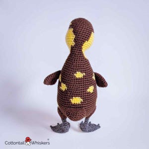 May include: A crocheted duckling toy featuring a dark brown body with scattered yellow spots, a yellow head, and dark gray webbed feet. The text Cottontail Whiskers with a small mushroom icon is printed in the bottom left corner.