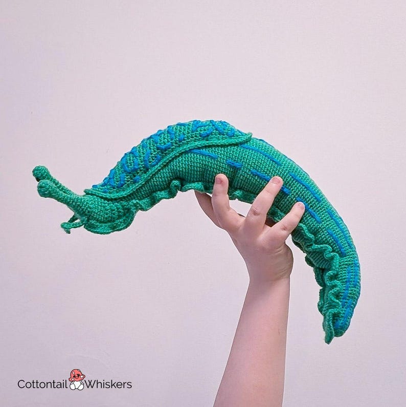 K&ouml;nnte beinhalten: Ein geh&auml;keltes gr&uuml;nes und blaues Pl&uuml;schtier in Form einer Schnecke. Die Schnecke ist zu einem C gekr&uuml;mmt und hat eine strukturierte Oberfl&auml;che.