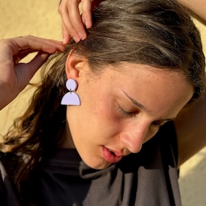 May include: A pair of lavender-colored geometric earrings. Each earring features a small circle above a semi-circle, connected by a thin silver wire. The earrings are worn on a person's ear.