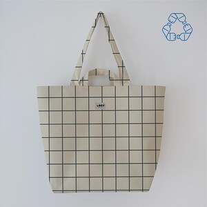 May include: A beige tote bag with a black grid pattern. The bag has two handles and a small label that reads "LORE". A recycling symbol is visible in the upper right corner. The bag is hanging against a white wall.