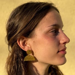 May include: A woman wearing gold glitter earrings with a semi-circle and circle design. The earrings are large and statement-making.