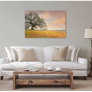 Op de afbeelding: Een witte bank met twee kussens staat in een woonkamer met een groot canvasprint van een eenzame boom in een veld vol wilde bloemen. De print hangt aan een lichtgrijze muur. Een houten salontafel staat voor de bank en een houten staande lamp staat rechts van de bank.