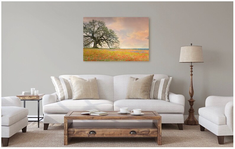 Op de afbeelding: Een witte bank met twee kussens staat in een woonkamer met een groot canvasprint van een eenzame boom in een veld vol wilde bloemen. De print hangt aan een lichtgrijze muur. Een houten salontafel staat voor de bank en een houten staande lamp staat rechts van de bank.