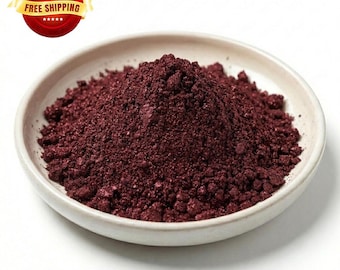 May include: A mound of deep burgundy, finely ground powder with a subtle metallic shimmer, contained within a small, shallow, off-white ceramic bowl. The texture of the powder appears granular and dense, filling the center of the round dish.