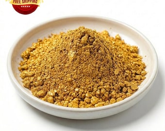 May include: A mound of metallic gold powder with a granular texture piled in the center of a shallow, round, off-white ceramic dish. The fine, shimmering particles create a dense, conical heap that fills the basin of the small, plain bowl.