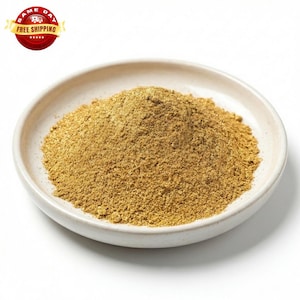 May include: A white ceramic bowl filled with a mound of fine, golden-brown powder. The powder has a granular texture and appears to be a spice or seasoning. The bowl is round with a slightly raised rim. The background is plain white.