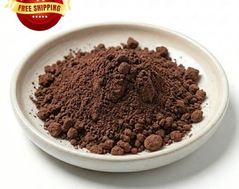 May include: A mound of dark brown, finely ground cocoa powder sits in the center of a shallow, off-white ceramic bowl. The powder has a textured, granular appearance with small clumps scattered throughout the pile, contrasting with the smooth bowl.