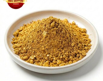 May include: A mound of metallic gold powder with a granular texture piled in the center of a shallow, round, off-white ceramic dish. The fine, shimmering particles create a dense, conical heap that fills the basin of the small, plain bowl.