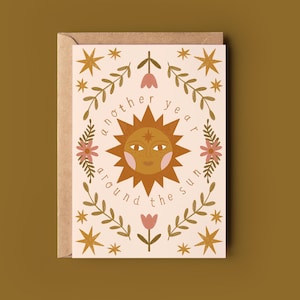 May include: A beige greeting card with a brown envelope. The card displays a sun illustration and the text "another year around the sun", encircled by floral and star designs in brown, pink, and gold.