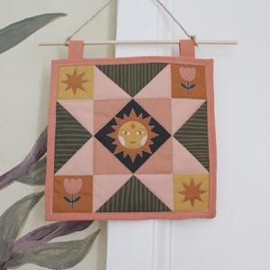 May include: A square quilted wall hanging featuring a central sun face motif surrounded by geometric patterns in shades of dusty pink, olive green, and mustard yellow. The design includes star and tulip icons, finished with a peach-coloured border.