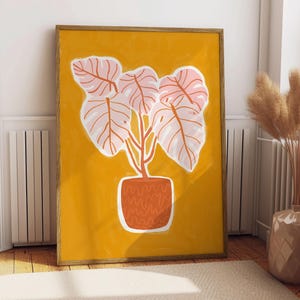 May include: A framed art print featuring a stylized illustration of a potted plant with large, light pink leaves and orange stems against a mustard yellow background. The pot is orange with a textured design. The artwork is displayed on a wall.