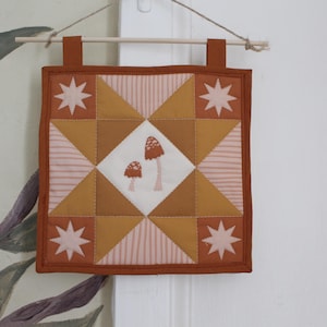 May include: A square fabric wall hanging featuring a central white diamond with two embroidered mushrooms. The quilt block design includes mustard yellow, rust orange, and pale pink striped fabric triangles with four white star motifs.