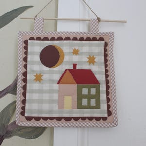 May include: A square fabric wall hanging featuring a house appliqué with a red roof, pink and green walls, and a yellow door. The scene includes a brown crescent moon and three yellow stars on a light grey and white gingham background with a scalloped border.