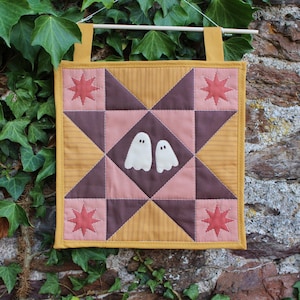 May include: A square quilted wall hanging featuring a central star pattern in mustard yellow, dark brown, and dusty pink. Two white ghost appliqués are centred on the quilt, with four red star motifs in the corners and two mustard fabric tabs.