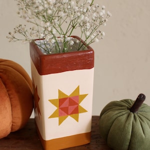 May include: A seasonal display featuring a cream-coloured rectangular vase with a starburst design, filled with white flowers. Either side of the vase are two plush pumpkins, one orange and one green, creating a cosy autumn aesthetic.