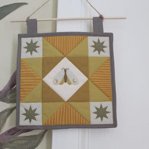 May include: A square quilted wall hanging featuring a central white diamond with a moth illustration. The quilt is composed of mustard yellow, olive green, and striped fabric blocks, accented with four eight-pointed stars in the corners, finished with a grey border and suspended from a wooden dowel.