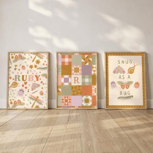May include: Three framed art prints with light wood frames. The prints feature whimsical designs with insects, flowers, and the text "RUBY" and "SNUG AS A BUG". The middle print has a patchwork quilt pattern with the letter "R".