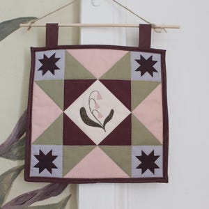 May include: A square fabric wall hanging featuring a patchwork quilt design with a central diamond containing an embroidered lily of the valley. The pattern includes dark maroon stars in the corners, light pink triangles, and sage green accents.
