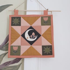 May include: A square fabric wall hanging featuring a central black diamond with a white crescent moon and mushroom graphic. The quilt block design includes pink, olive green, and mustard yellow geometric patterns, stars, and tulip motifs.