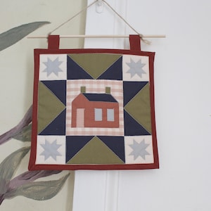 May include: A square fabric wall hanging featuring a central block with a house illustration on a light pink gingham background, surrounded by navy blue and olive green triangles and four light grey stars, all framed by a dark red border.