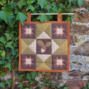 May include: A square quilted wall hanging featuring a patchwork design with brown and olive green fabric squares. The pattern includes four star motifs in the corners and a central square displaying an appliqué of an acorn with two oak leaves.