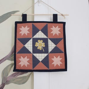 May include: A square fabric wall hanging featuring a patchwork star design in muted terracotta, navy blue, and light grey gingham. The centre square displays a yellow four-leaf clover graphic. It is suspended from a wooden dowel with twine.