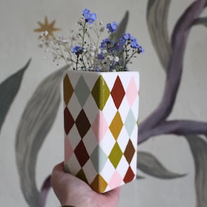 May include: A rectangular white vase with a harlequin pattern in shades of brown, pink, green, and yellow. The vase holds a small bouquet of blue and white flowers. The vase is held in front of a wall with a floral design.