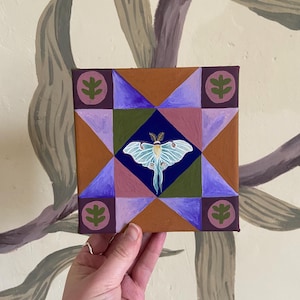 May include: Square canvas art with a geometric design and a central moth illustration. The artwork uses brown, purple, green, and blue. The moth is light blue with yellow accents. The corners of the canvas have a leaf design.
